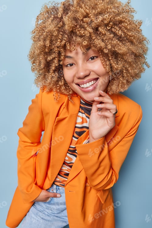 Preview: Happy curly haired young European woman smiles broadly dressed in fashionable orange jacket and