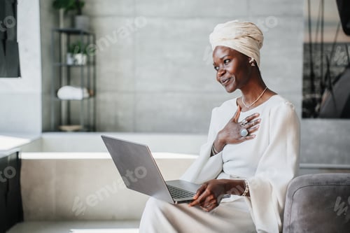 Vista previa: Mujer africana agradecida con turbante blanco, vestido sentado en casa hace una videollamada con el ordenador portátil