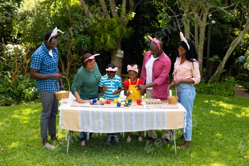 Preview: Happy african american multigenerational family in bunny ears painting easter eggs in backyard