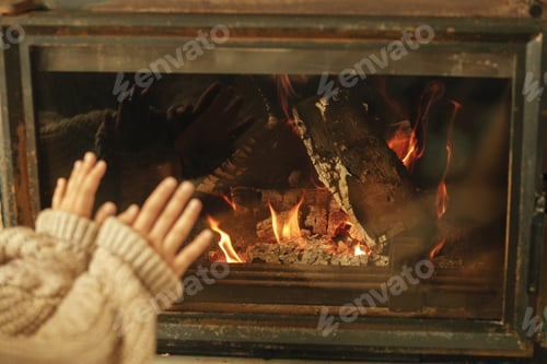 Preview: Fireplace heating in winter. Woman in cozy sweater warming up hands at fireplace in rustic room