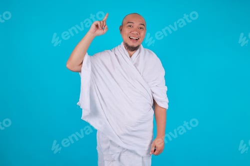 Preview: Happy Asian Man in White Ihram Clothing Pointing up Blue Background