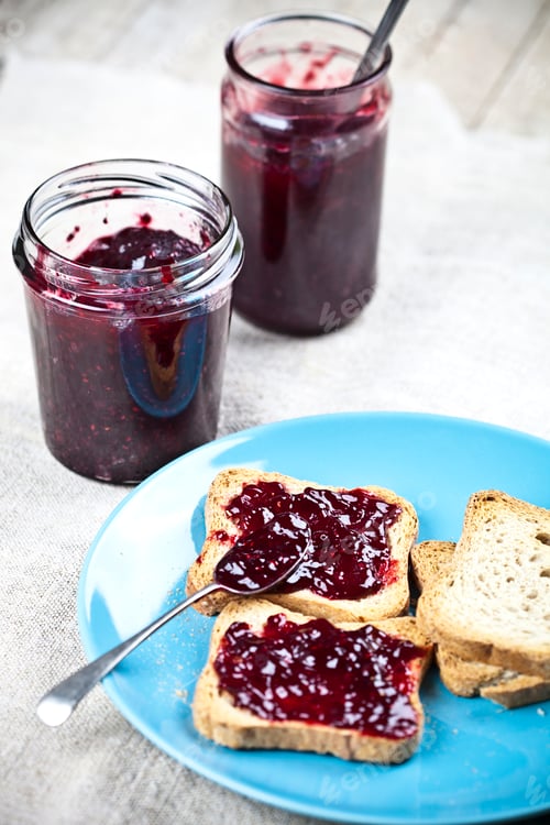 Preview: Fresh cereal bread slices on blue ceramic plate, homemade cherry and wild berries jam in jars.
