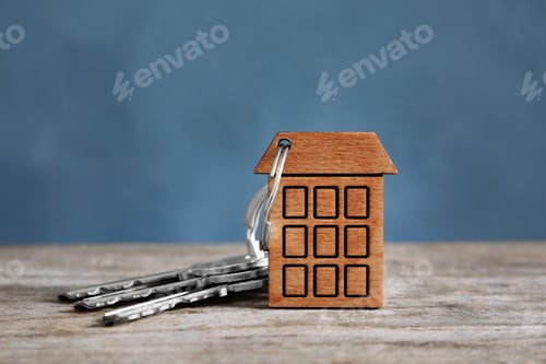 Preview: House keys with trinket on wooden table against color background. Space for text