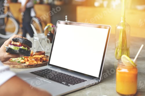 Preview: Male freelancer eating burger during lunch at cafe, sitting at table with open laptop and fresh juic
