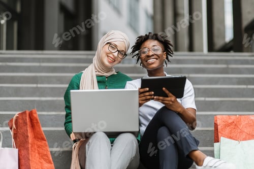 Preview: African and Muslim women sitting on the steps of mall with laptop and tablet and shopping online.