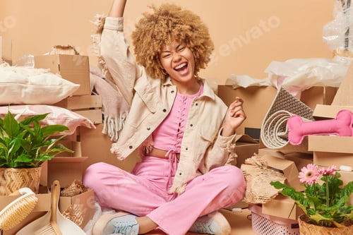 Preview: Positive curly haired woman sits crossed legs on floor with various household items around exclaims