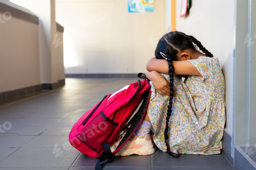 Preview: Sad biracial schoolgirl with school bag sitting at wall at elementary school corridor, copy space