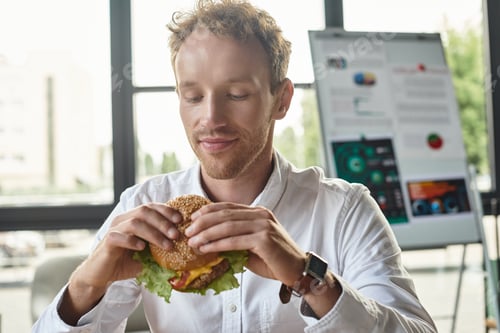 Preview: A focused redhead businessman enjoys a tasty burger in his modern office at lunch