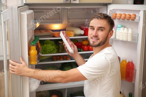 Preview: Young man taking meat from refrigerator in kitchen