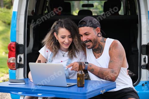 Preview: Young tattooed couple smile and see something in the laptop near by the van.