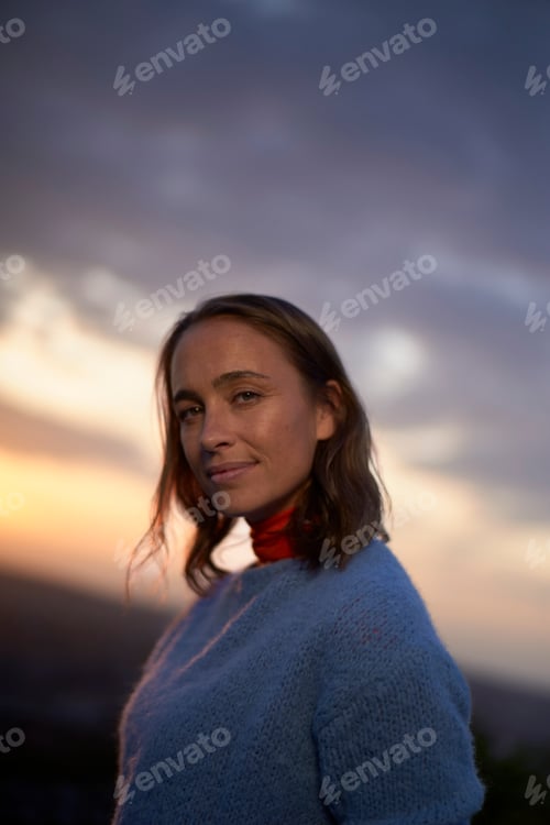 Preview: Smiling woman in a blue sweater with a red scarf standing against a sunset sky background.