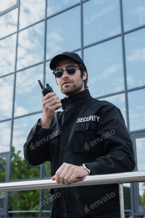 Preview: security man in sunglasses and black uniform talking on walkie talkie outdoors