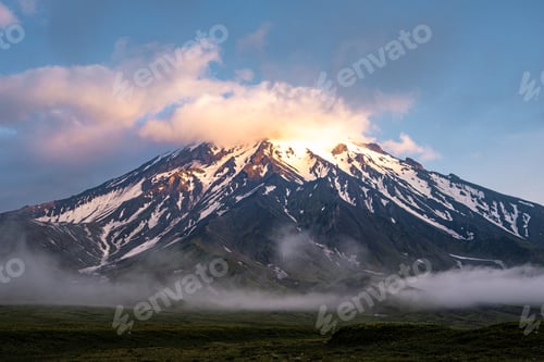 Preview: A smoking snow-covered volcano in the clouds illuminated by the sun. Volcanic activity.
