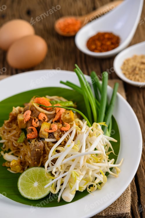 Preview: Pad Thai in a white plate with lemon, eggs and seasoning on a wooden table.
