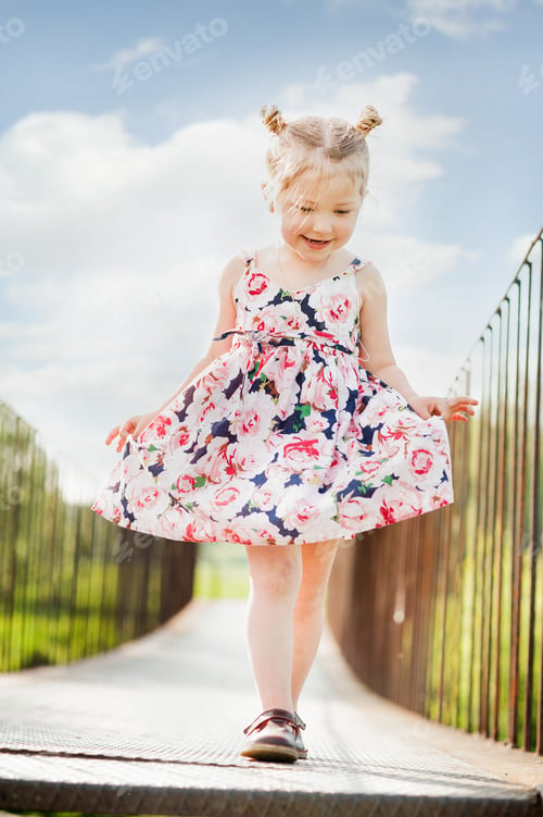 Preview: Happy girl in a summer dress with pigtails plays on a wooden bridge