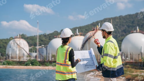 Preview: Two engineers in safety gear with blueprints discussing work in front of industrial storage tanks.