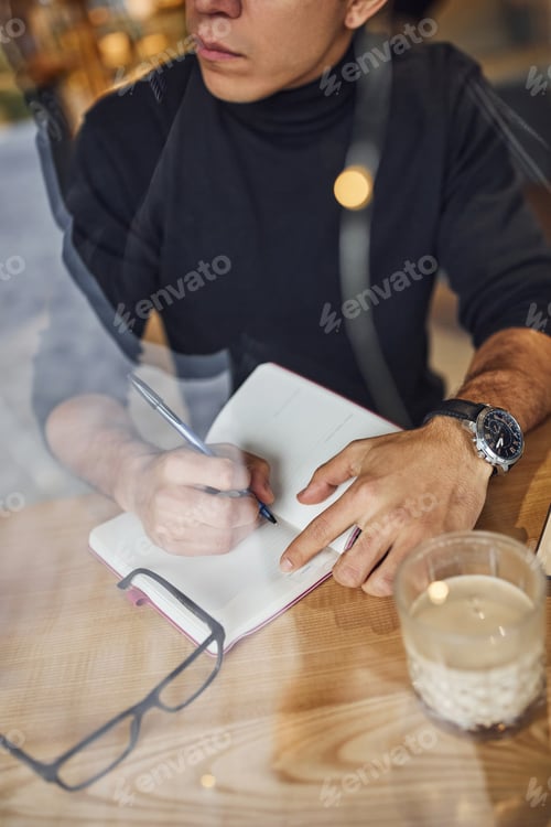 Preview: Young male writer with black curly hair sitting indoors in cafe with pen and notepad
