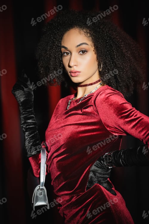 Preview: Portrait of trendy african american woman in dress posing with handbag on burgundy drapery