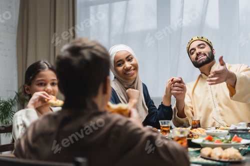 Preview: Smiling muslim parents holding hands near blurred kids and food during ramadan at home