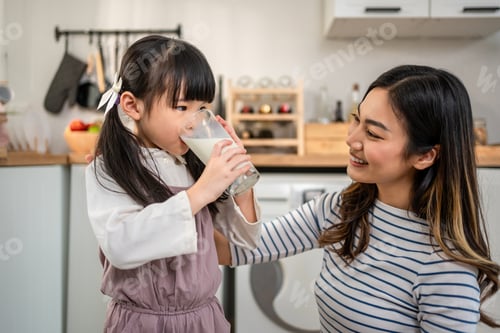 Preview: Asian little daughter drinking a glass of milk with mother in kitchen.