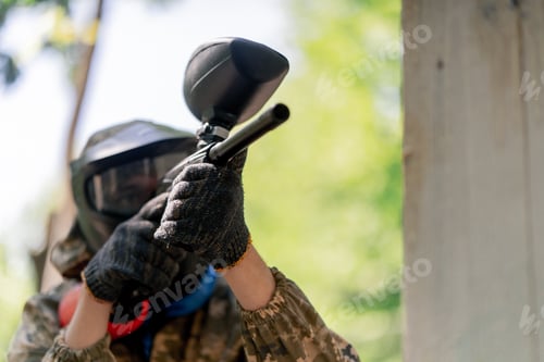 Preview: close up in the wild near tree structures player in a pintball uniform holds a pistol with a red