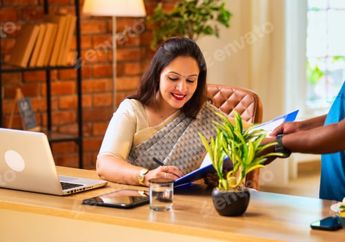 Preview: Indian woman in saree signs file while working on laptop at cozy corporate desk