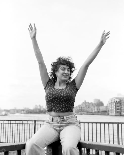 Preview: Young woman raising arms by river in monochrome analog medium format film photography