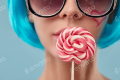 Preview: Croped shot of young funky girl in blue wig and eyeglasses, holding lollipop candy, looking aside