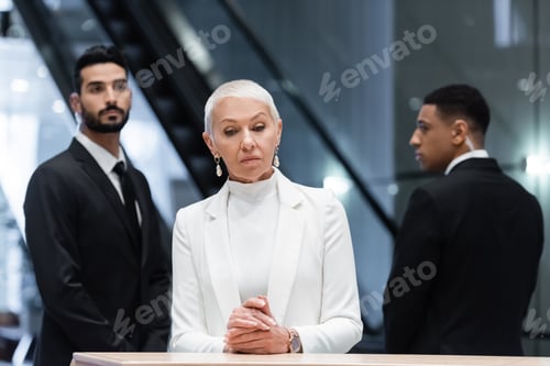 Preview: stylish senior business lady standing at hotel reception near blurred interracial bodyguards