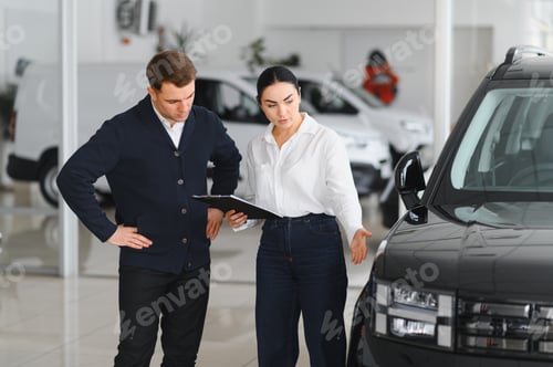 Preview: Friendly female car dealer standing at fancy car showroom and having consultations with a customer