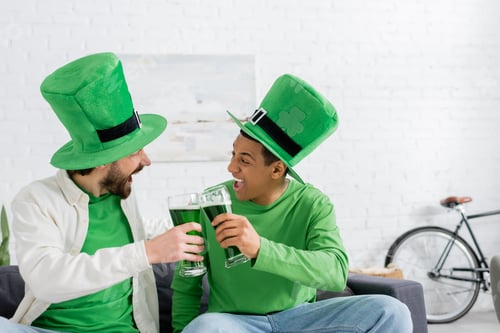 Preview: Excited multiethnic friends in green hats clinking beer during saint patrick day at home