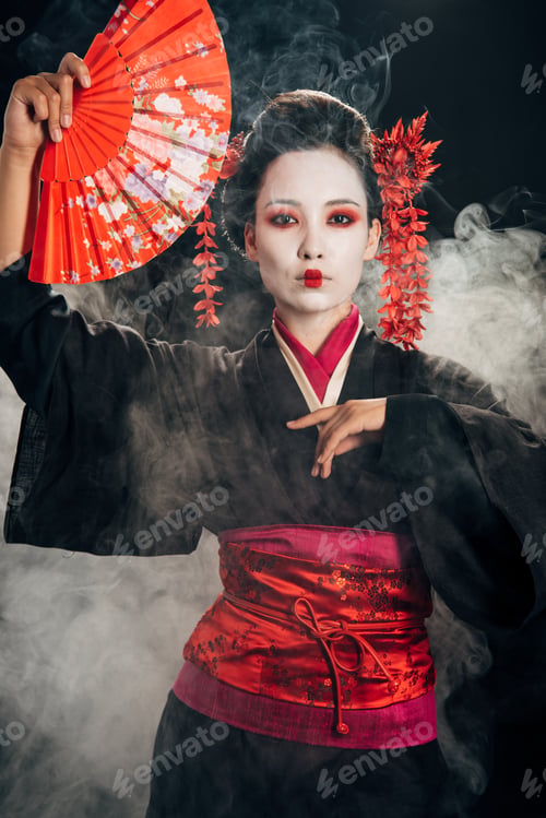 Preview: beautiful geisha in black kimono with flowers in hair holding hand fan and gesturing in smoke