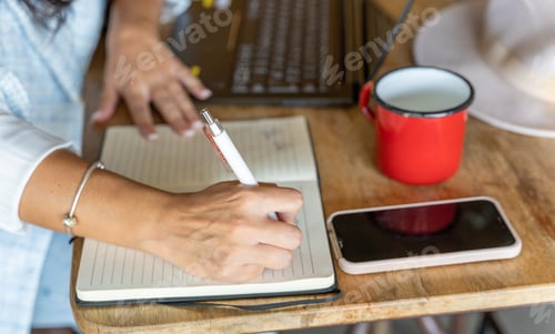 Preview: Close-up view of a woman writing on a notebook while working with a laptop in a coffee shop.