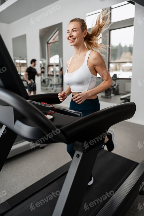 Preview: Young woman jogging on treadmill in modern gym, training for health and fitness goals