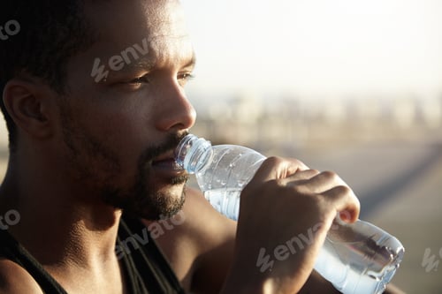 Preview: Attractive young dark skinned sportsman with short beard drinking water from bottle looking far away