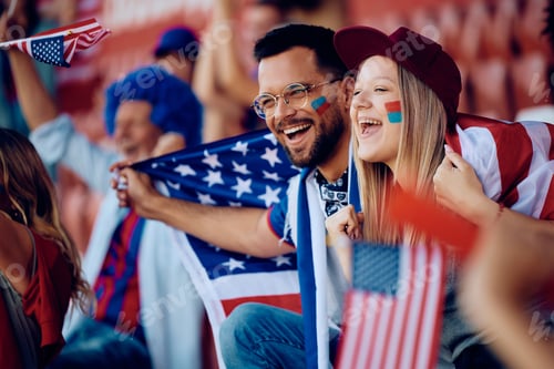 Preview: Happy couple of sports fans wrapped in USA national flag watching a game at the stadium.