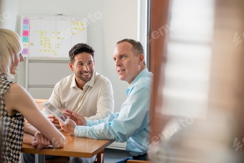 Preview: Three business colleagues in an office talking around a table and looking at a digital tablet.