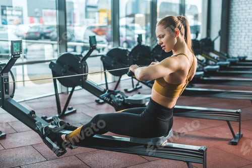 Preview: Young sportswoman doing exercise on rowing machine in sports center