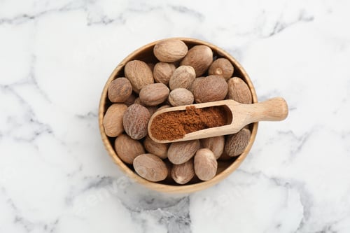 Preview: Scoop with nutmeg powder and seeds in bowl on white marble table, top view