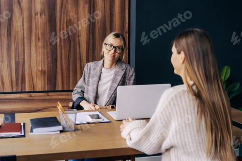 Preview: Two young modern business women smiling talking while sitting at the desk in the office.