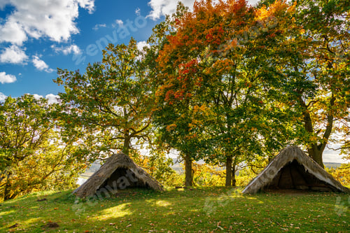 Preview: Two rustic shelters with thatched roofs on a grassy hillside under the canopy of autumn trees