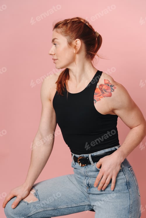 Preview: Modern sporty muscular woman with red hair in t-shirt and jeans posing on pink background