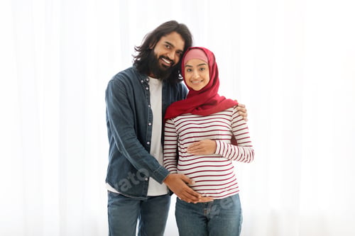 Preview: Young Smiling Pregnant Arabic Family Standing Near Window At Home