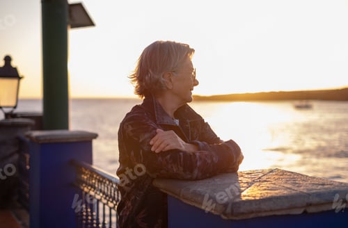 Preview: Handsome senior woman in denim jacket looking at sea while the sun goes down.