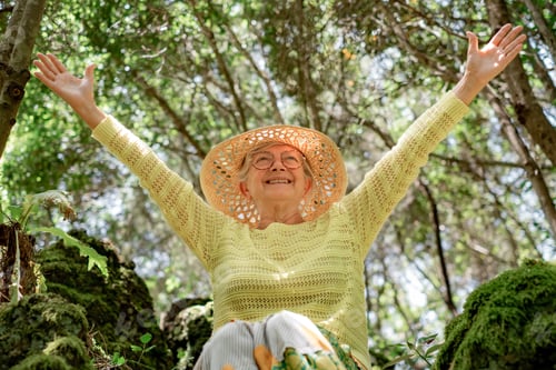 Preview: Smiling happy mature senior woman in outdoors excursion in the forest sitting with outstretched arms