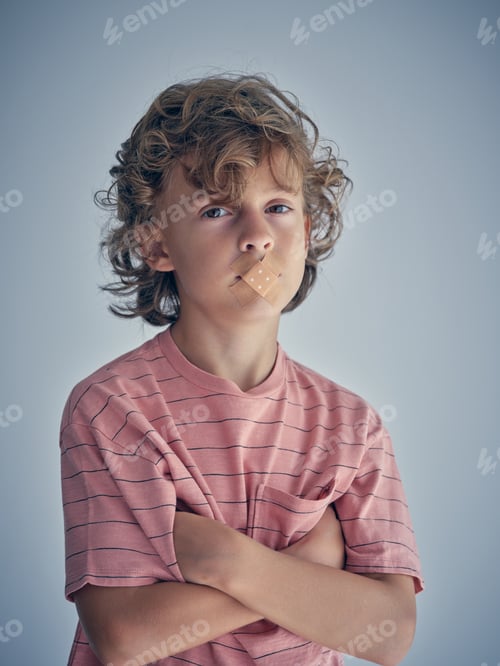Preview: Upset boy with plasters covering lips on gray backdrop