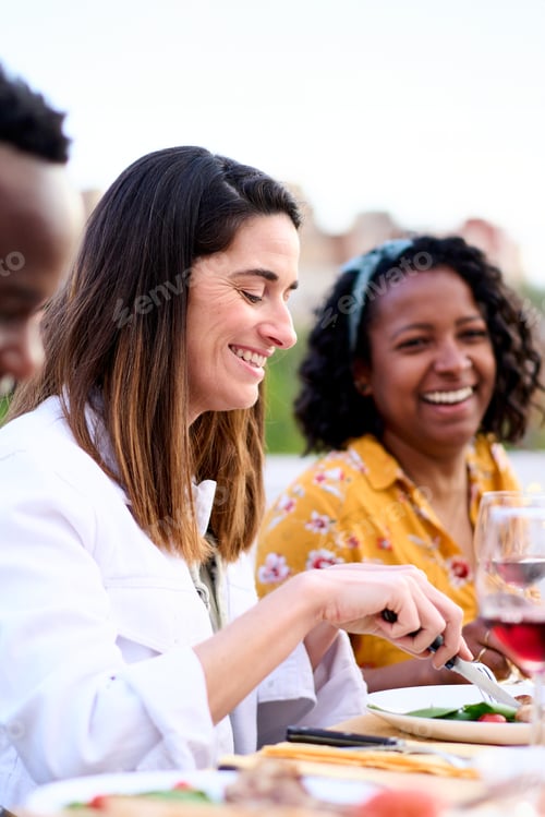 Preview: Side view of smiling Caucasian woman eating food sitting at table with friends unfocused on rooftop.