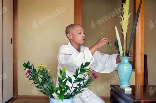 Preview: Buddhist monk with shaved head wearing white robe kneeling on floor, arranging flowers in blue vase.
