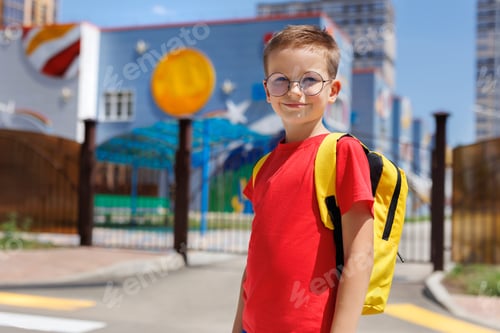 Preview: Boy in bright clothes, glasses and with a backpack poses against the background of a modern school.