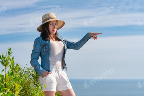 Preview: Hipster woman looking at camera, pointing hand to side, standing on background of blue sky and sea